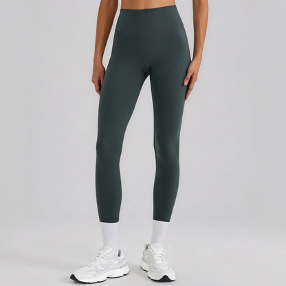 A model from the waist down wearing high-waisted, full-length athletic leggings in a dark forest green color, paired with white socks and white sneakers, against a light gray studio background.