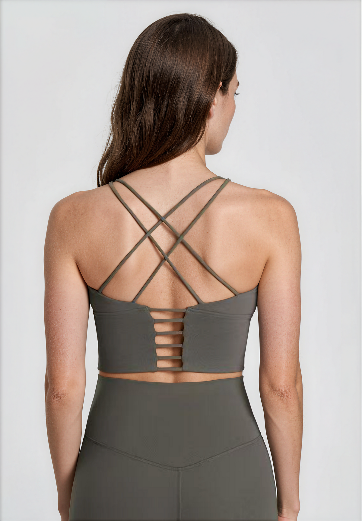 A woman shown from the back wearing a dark khaki athletic top with crisscrossing straps and a cutout back design, paired with matching high-waisted leggings against a light gray studio background.