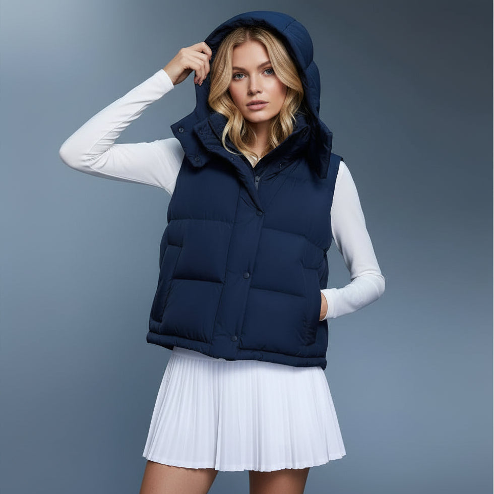 Woman wearing a navy puffer vest over a white long-sleeve shirt and pleated skirt on a gray background