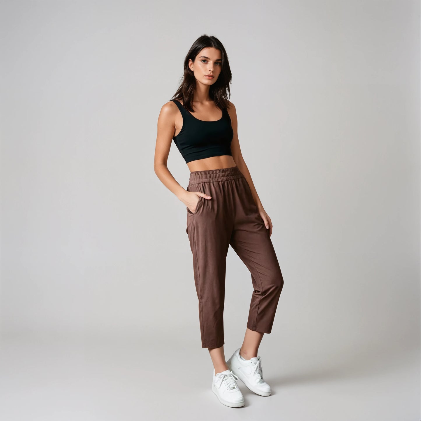Woman wearing a black tank top and coffee brown joggers standing against a concrete wall.