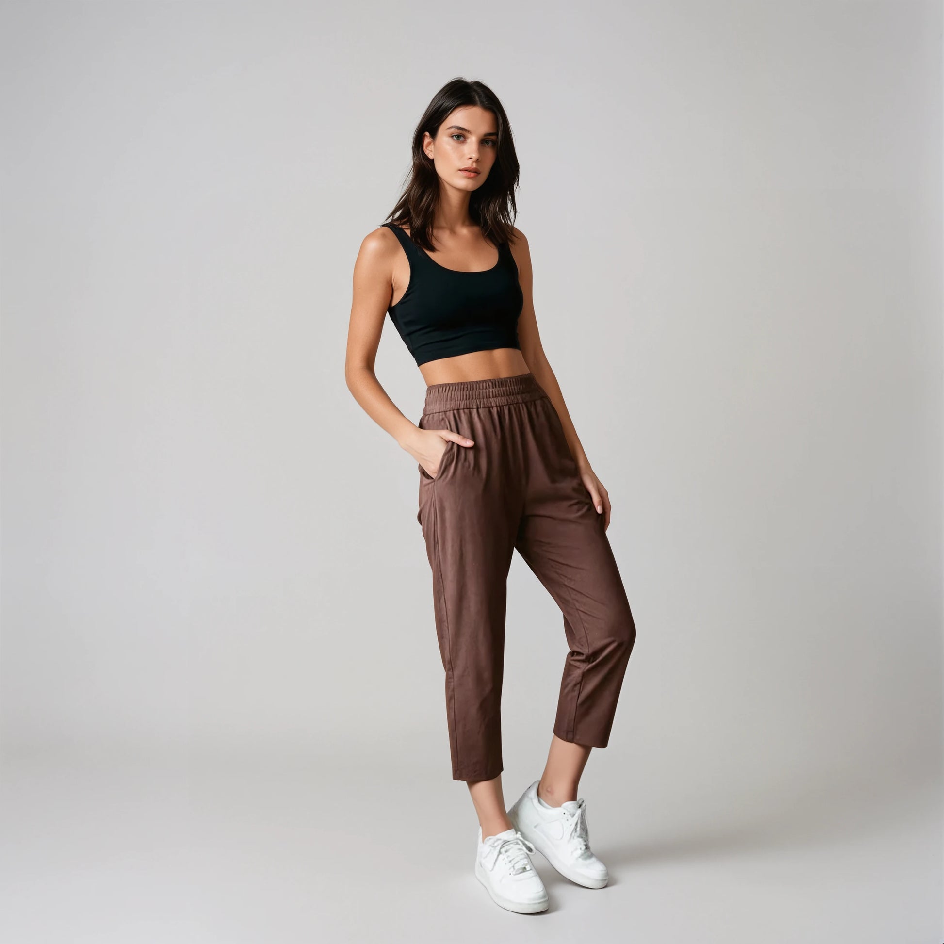 Woman wearing a black tank top and coffee brown joggers standing against a concrete wall.