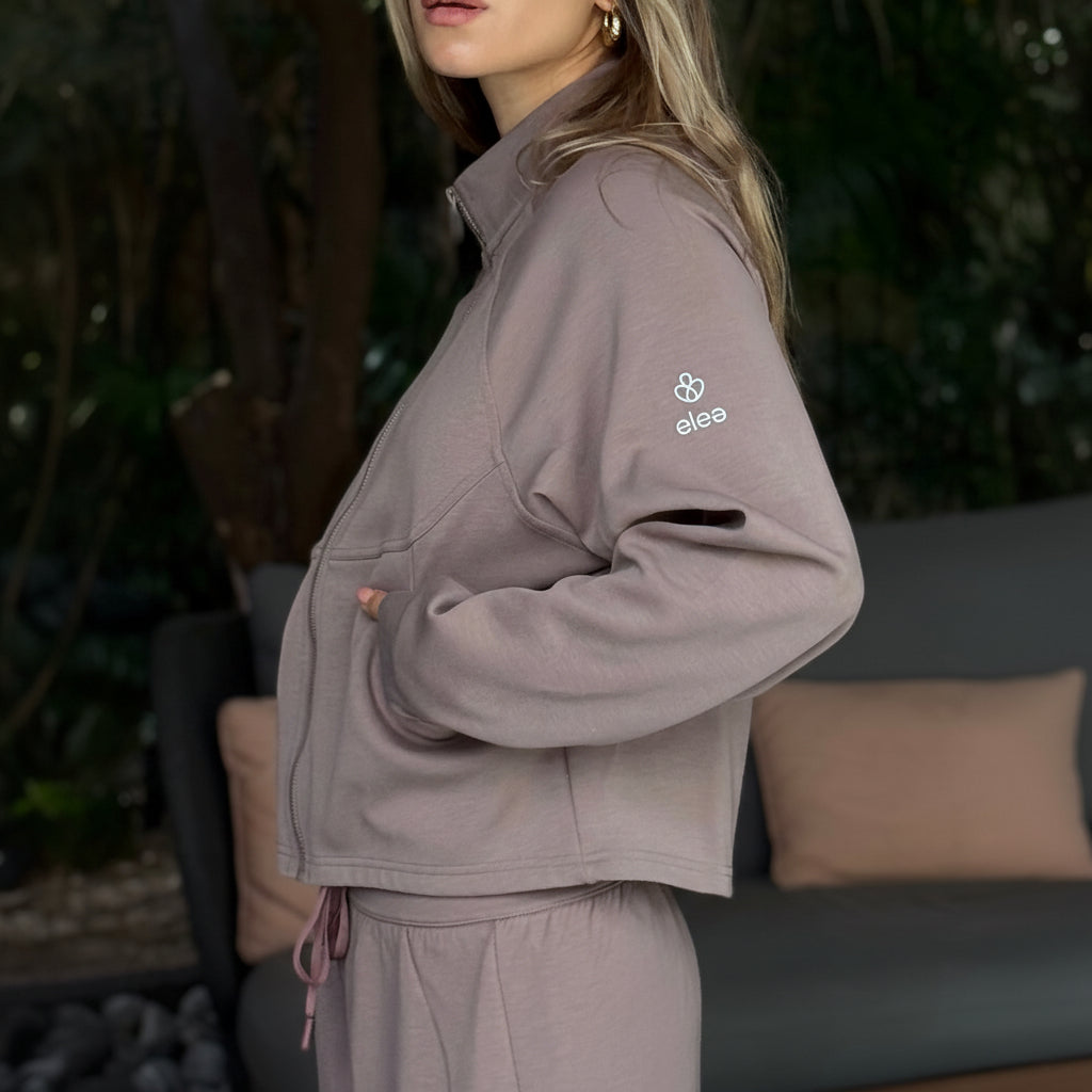 Roni wearing a taupe tracksuit with an elea logo on the sleeve, standing outdoors.
