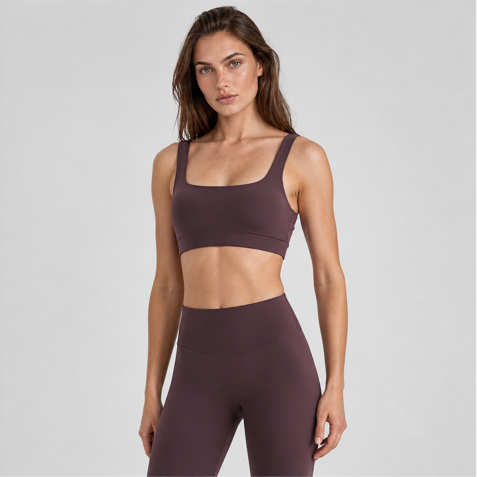 A woman modeling a matching athletic set consisting of a scoop-neck sports bra and high-waisted leggings in a solid dark plum purple color, set against a plain light gray studio background.